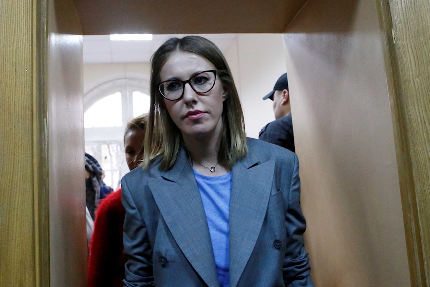 A blonde Ksenia Sobchak walks towards the camera wearing thick frame glasses and looking to the left.