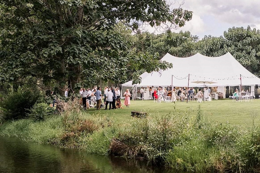People gathered for a wedding around a large white medieval shaped tent which is positioned by a creek