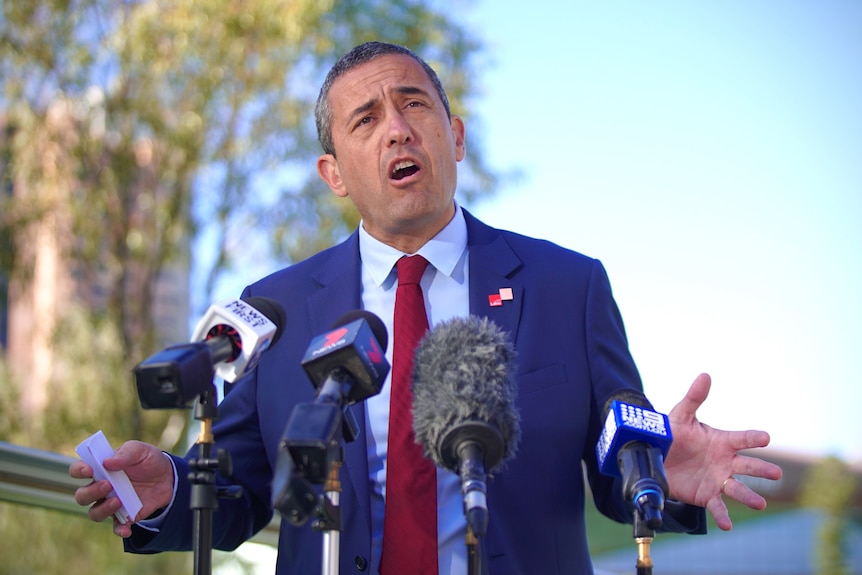 A man wearing a suit and tie gesticulates while speaking to microphones