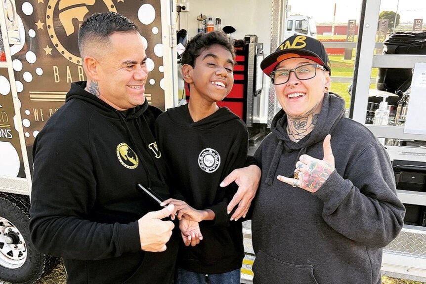 Three people standing in front of a mobile barber shop smiling, one is a 12 year old boy in the middle with freshly cut hair.