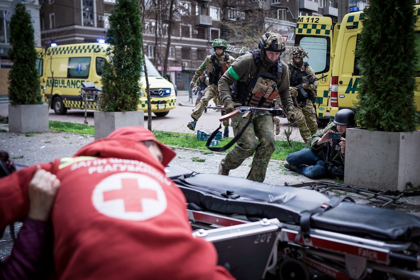 一名身穿红十字夹克的男子蹲在地上，军队从他身边跑过 