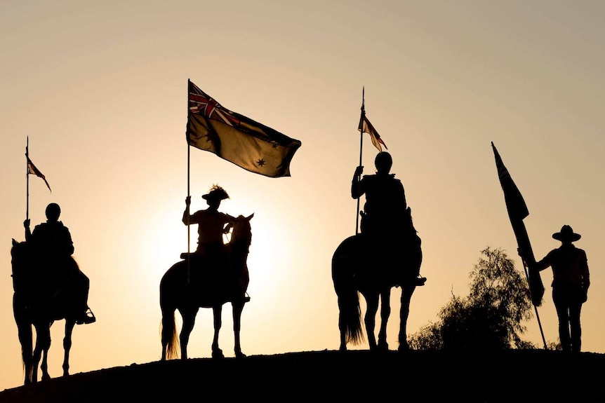 Four mounted horsemen carrying Australian flags are silhouetted against a setting sun