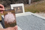 The hands of a woman holding a photo of a dirt grave and headstone with a grave behind it
