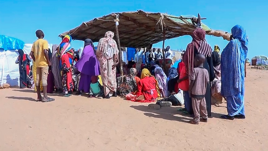 A group of refugees wearing colourful clothing gather under a shelter protecting them from the sun