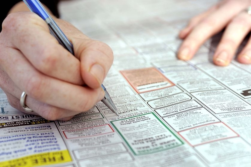 A close-up shot of a newspaper page showing jobs advertisements with a hand holding a pen above it.