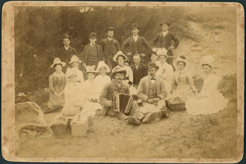 A sepia-toned faded picture of people in 1880s suits and dresses at a beach picnic posing seriously for the camera