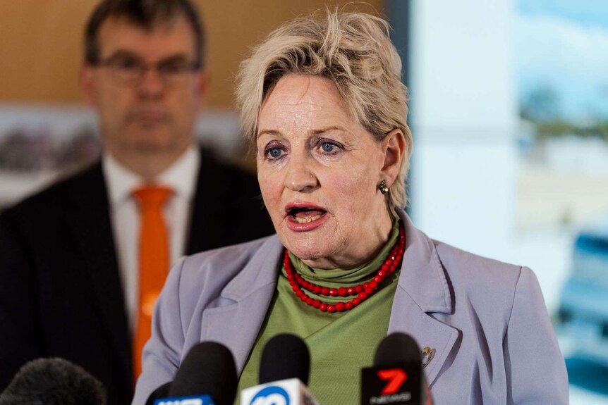 a woman with short, light hair and wearng a suit jacket speaking at a news conference