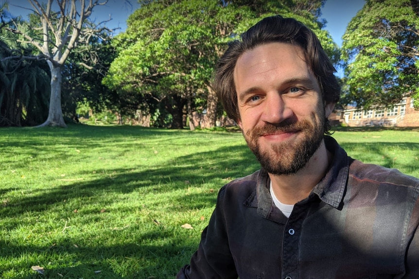 Brown-haired man with beard and dark plaid shirt sits in grassy park