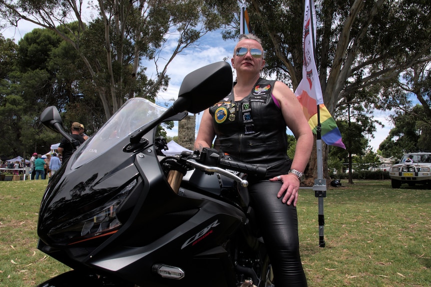 A woman with sunglasses on and half her head shaved sits on a motorbike with her hand on her thigh.