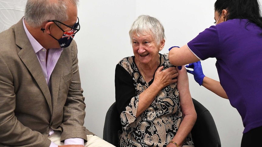 Jane Malysiak receives a needle jab in her arm from a nurse while Prime Minister Scott Morrison sits next to her and watches on.