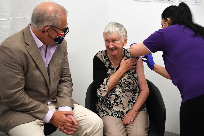 A middle-aged man sits beside an elderly woman receiving a needle in the arm