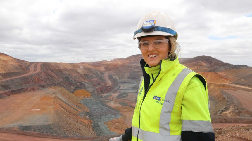Girl in the foreground left in high vis gear, helmet glasses, overlooking a mine pit that has step layers of mining