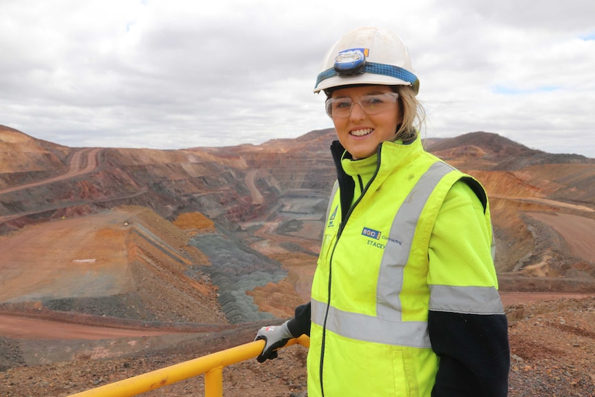 Girl in the foreground left in high vis gear, helmet glasses, overlooking a mine pit that has step layers of mining