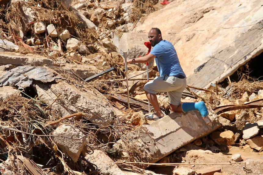 A man with a leg cast and crutches kneels down in a pile of rubble and debris