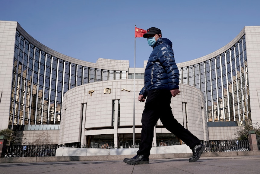 Ein Mann mit Gesichtsmaske und blauer Daunenjacke geht an einem hohen Gebäude vorbei, an dessen Stange eine chinesische Flagge weht.