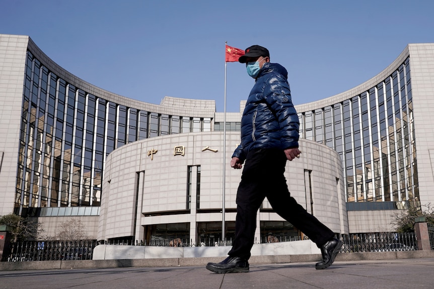 A man wearing a face mask and blue puffer jacket walks past a large building with a Chinese flag flying on a flagpole.