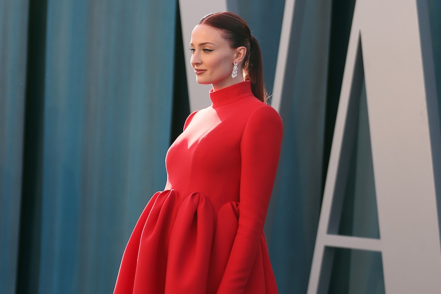 Sophie Turner smiles while posing in a red dress and dangly earrings.