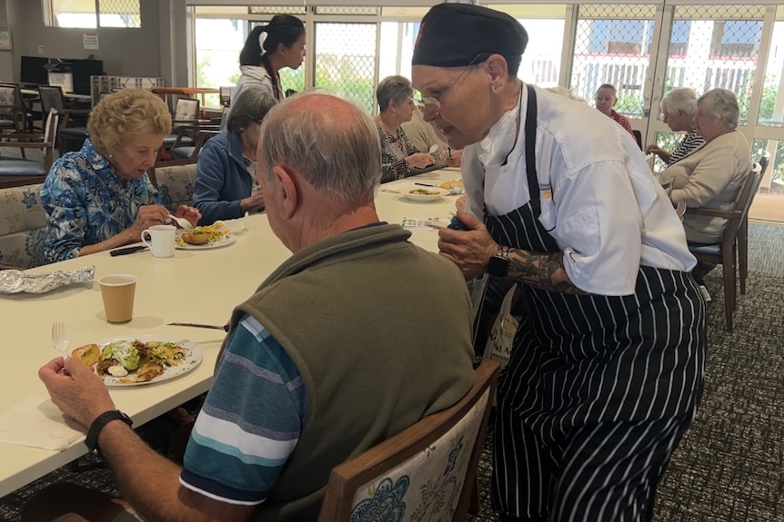 A chef leans down to talk to an elderly gentlemen eating a meal at the table