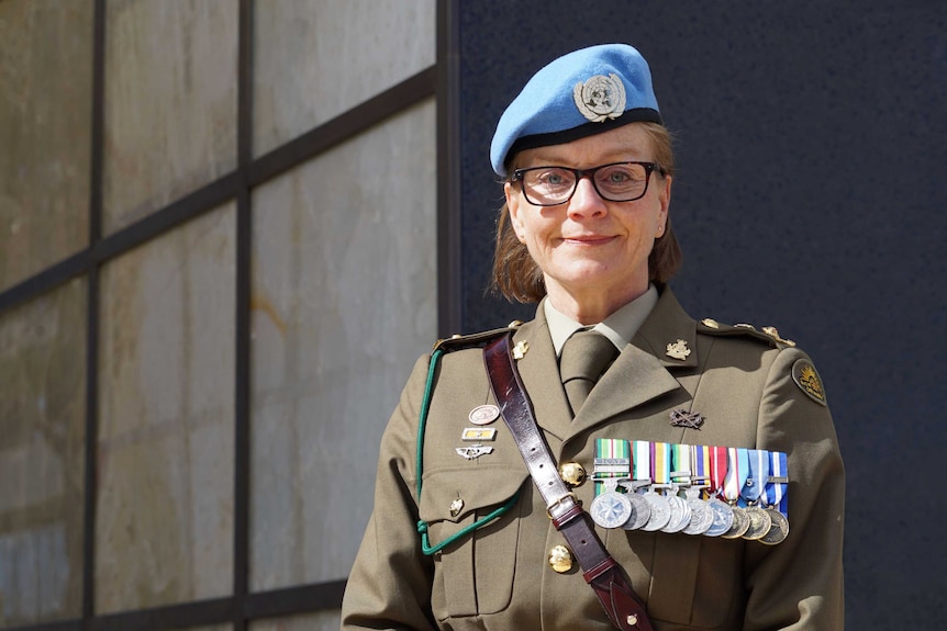 A woman wearing medals stands in front of a memorial.