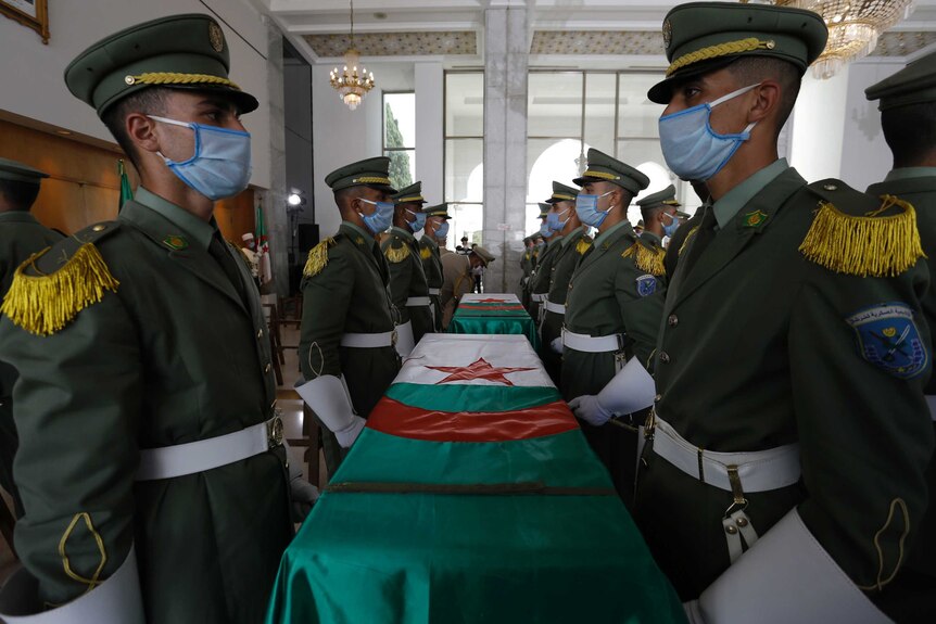 Soldiers in dress uniforms wearing face masks stand in ranks alongside coffins draped with red, white and green flags.