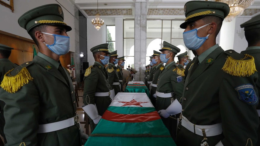 Soldiers in dress uniforms wearing face masks stand in ranks alongside coffins draped with red, white and green flags.