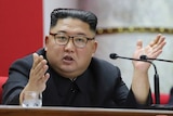 Kim Jong-un sits in front of two microphones with slicked back hair and gestures, wearing tortoiseshell glasses.