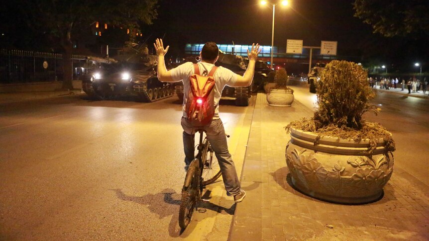 A man sits on a bike with his hands in the air, while in front of him are two military tanks on a road.