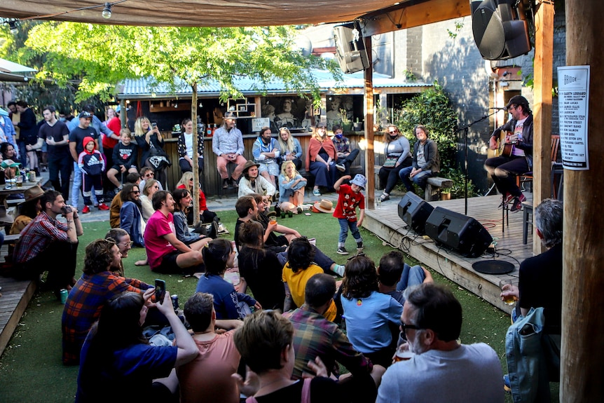 Small sunny outdoor music event with seated audience looking up at male guitarist while a child dances at foot of a small stage 