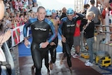 A man, George Skoufis, running in a wetsuit with ahead of other competitors and spectators watching from barricades.