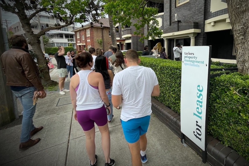 Dozens of people of all ages line up outside a "For Lease" sign at a Sydney apartment building.