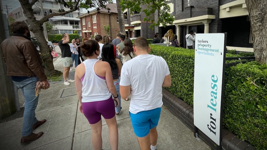 Dozens of people of all ages line up outside a "For Lease" sign at a Sydney apartment building.