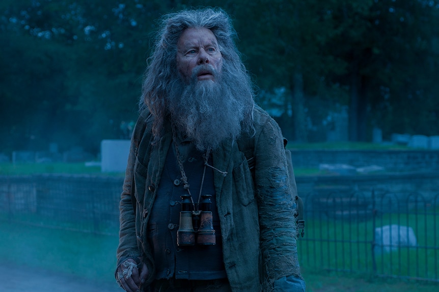 A man with brilliant silver hair and beard with awed expression and wearing tattered coat and binoculars stands near graveyard.