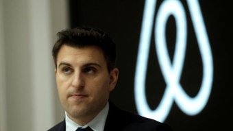 Brian Chesky looks up as he stands in front of a black and white Airbnb logo. He wears a black suit and white shirt.