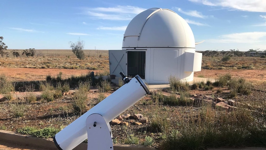 A round-roofed observatory can be seen behind a telescope pointed at the sky in broad daylight on brown dirt.
