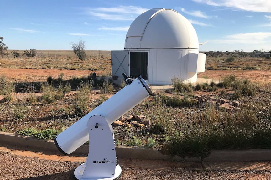 A round-roofed observatory can be seen behind a telescope pointed at the sky in broad daylight on brown dirt.