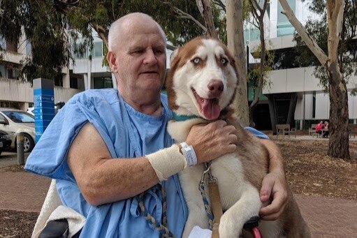 Rennie sits in front of a hospital wearing a blue gown. He has his arms around a large brown and white dog.