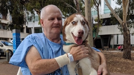 Rennie sits in front of a hospital wearing a blue gown. He has his arms around a large brown and white dog.
