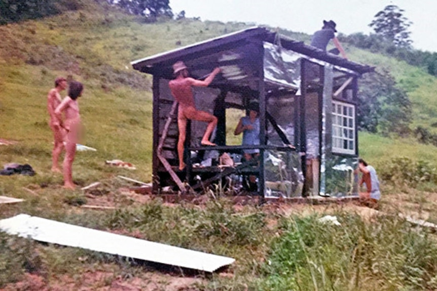 Naked and barely clothed men and women working together on a building project on a green hillside.