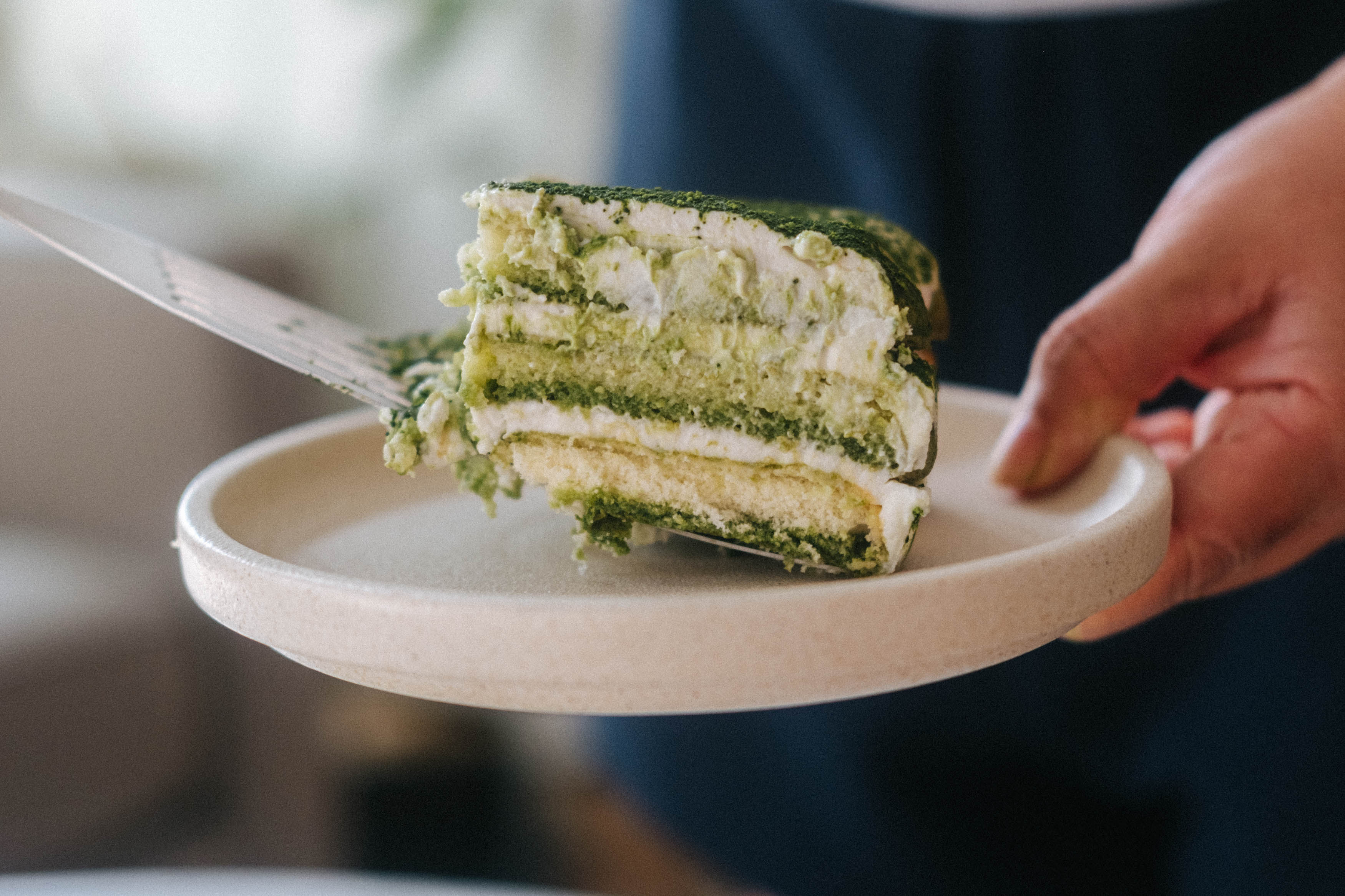 A slice of matcha tiramisu being served onto a white plate using a spatula. The layers of biscuit, cream and matcha visible.