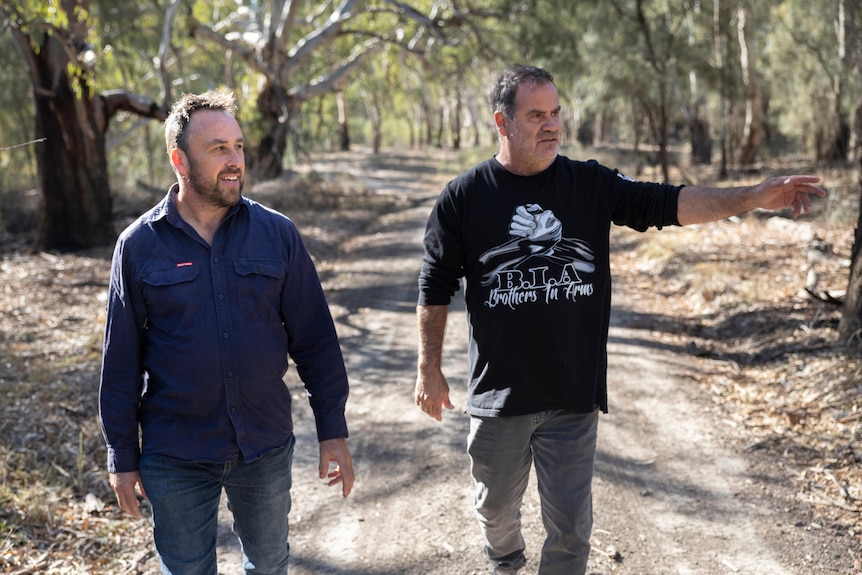 two men walk down a bush track, one is indicating to right of the frame and the other is looking at where he's pointing