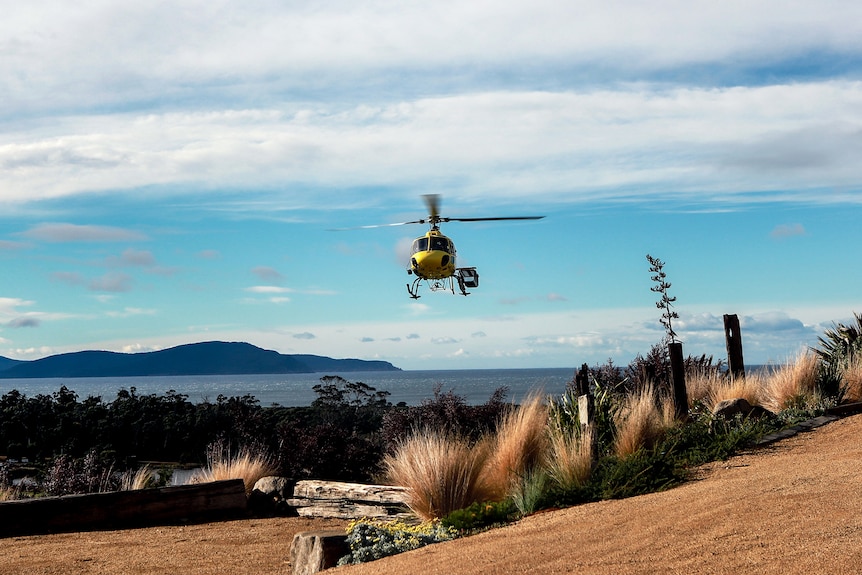 A yellow helicopter hovers above a gravel path with coastline and blue sky in background