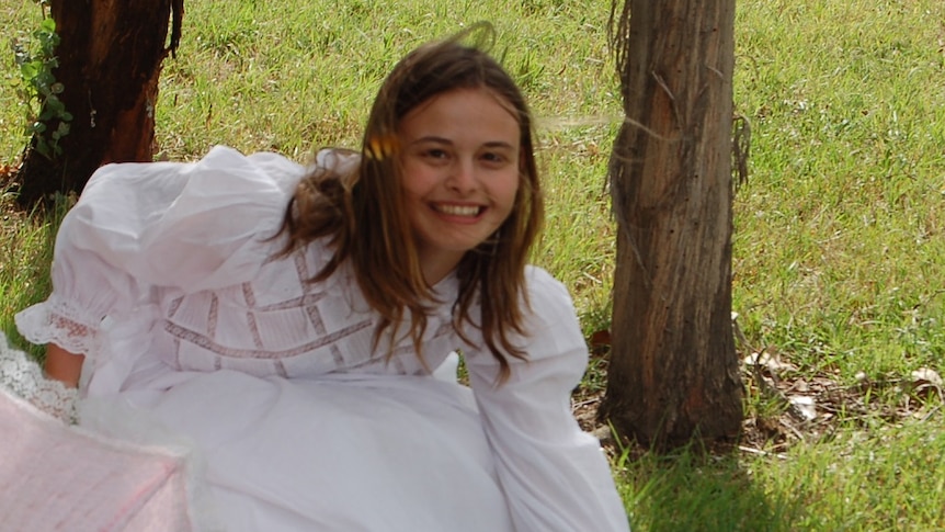 A young girl wearing a white dress sits on green grass and smiles at the camera.