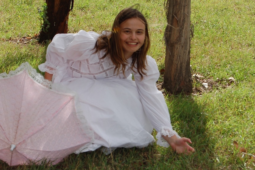A young girl wearing a white dress sits on green grass and smiles at the camera.