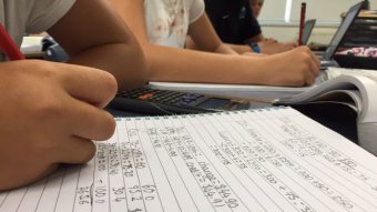 A close-up photograph of a student's hands writing out maths problems.