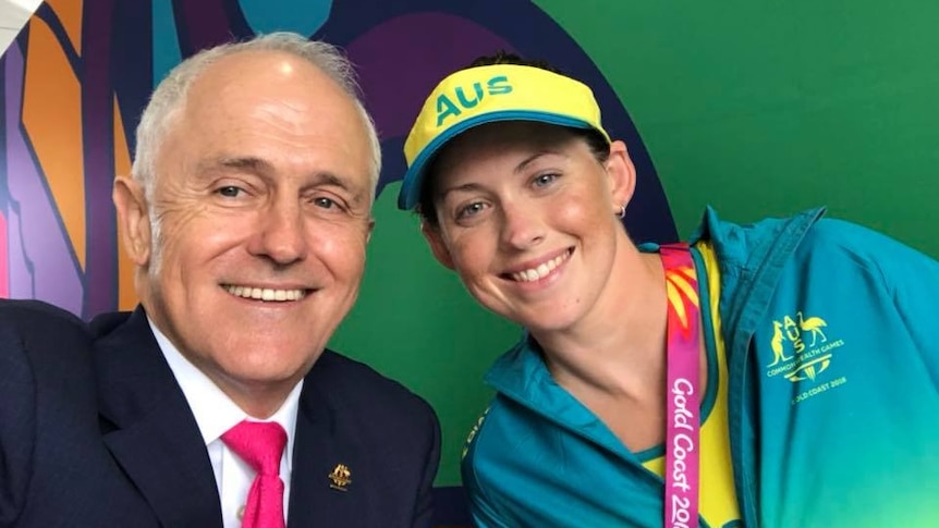 A man wearing a suit and a woman wearing a green tracksuit, yellow visor and medal smile at the camera.