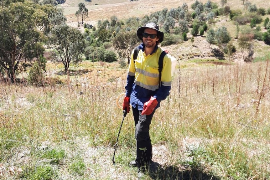 A man wearing high visibility clothing holding a spray gun in a paddock with rolling hills in the background.
