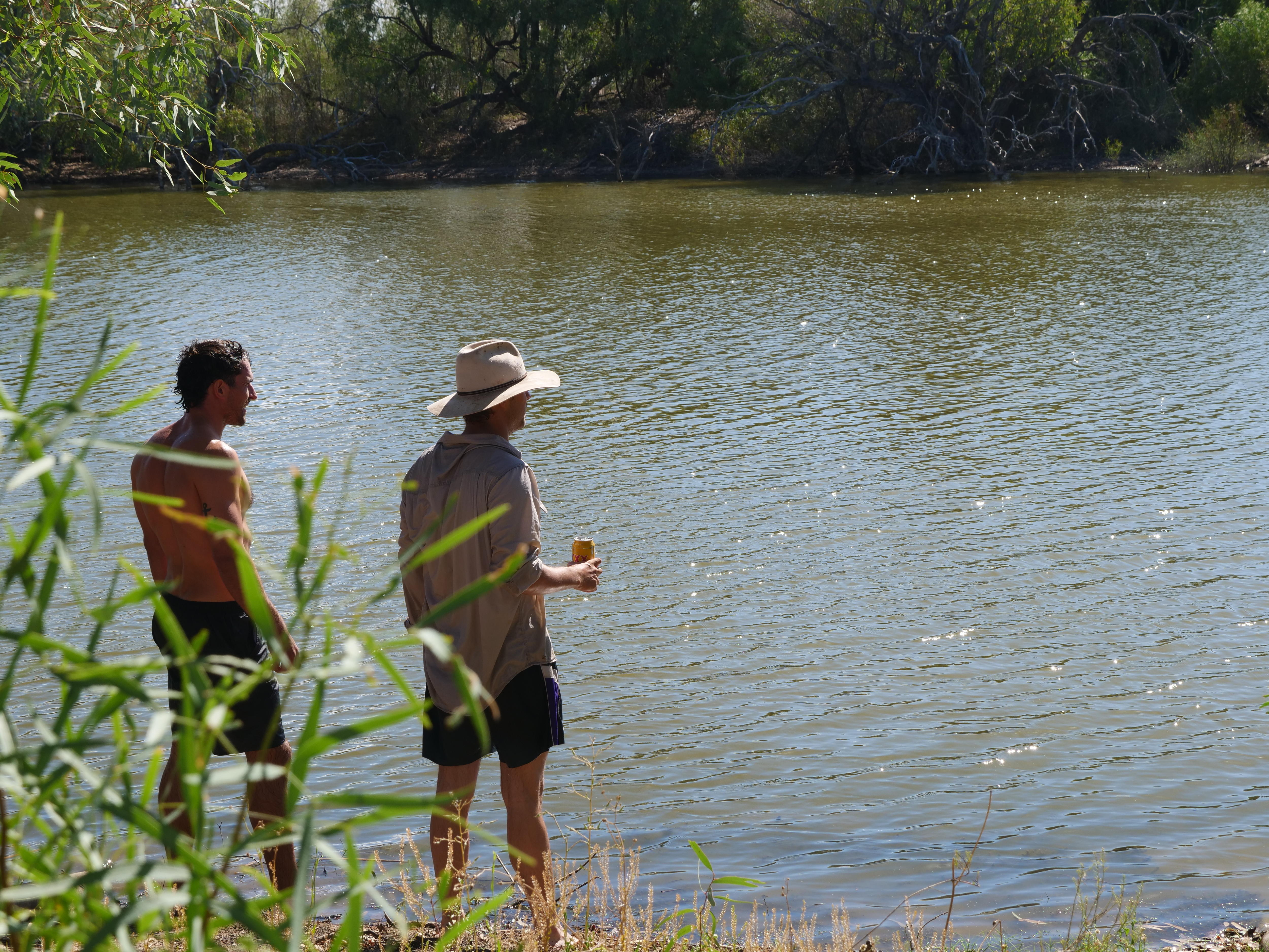 Dois jovens estão em frente a um rio marrom brilhante segurando uma cerveja. 