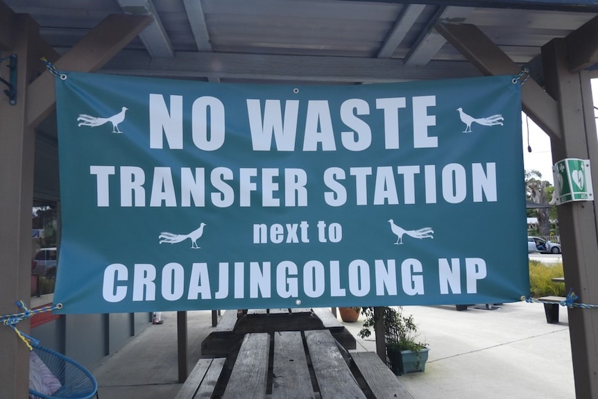 A green sign with white text and peacocks says "no waste transfer station next to corajingolong NP"