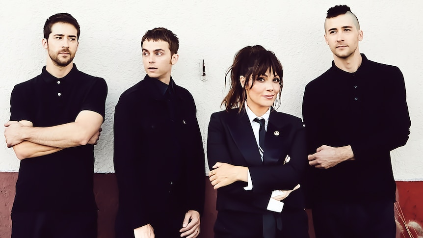 Three men dressed in black and a woman wearing a suit stand in front of an off-white wall