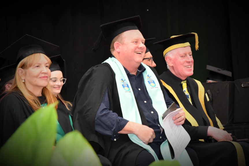 A man in a university cap and gown laughing sitting between a man and a woman